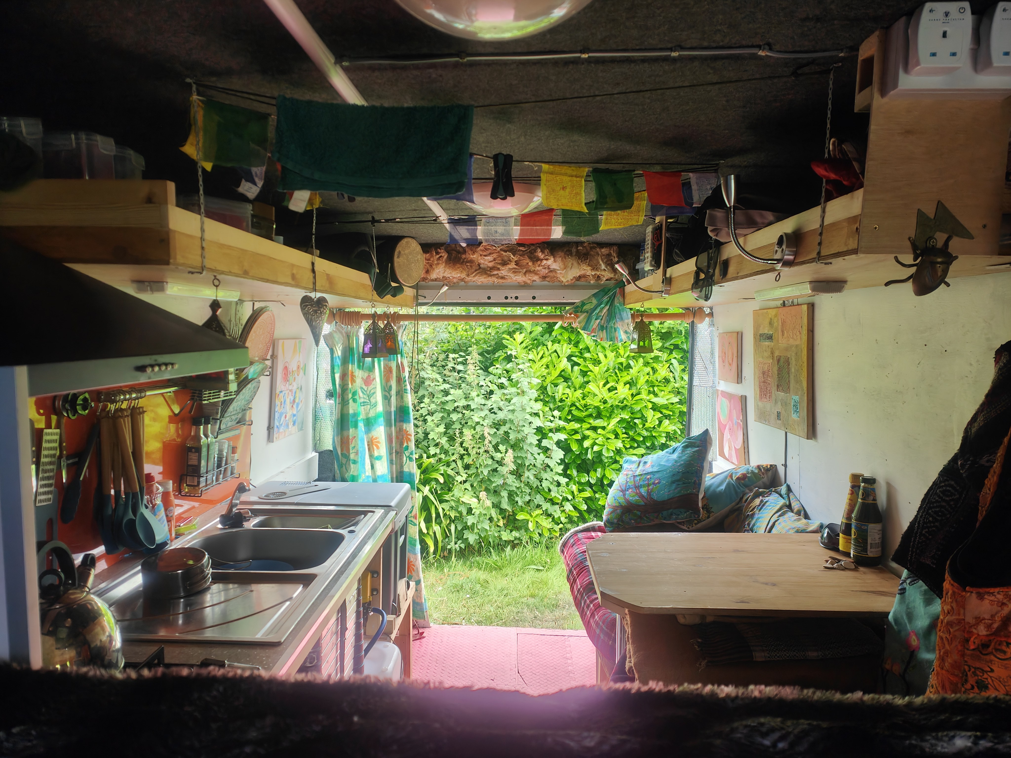 Looking out through the back doors of the Mercedes Vario motorhome conversion — handmade kitchen, prayer flags, and Devon greenery beyond